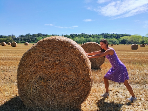 A woman pushing a large hay bale in a field, seeking marketing support.